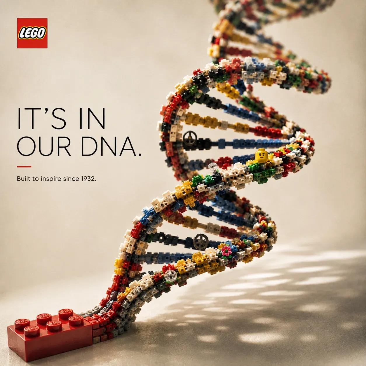 LEGO double-helix DNA sculpture built from multicolored bricks rising off a red LEGO baseplate, headline IT IS IN OUR DNA — built to inspire since 1932.